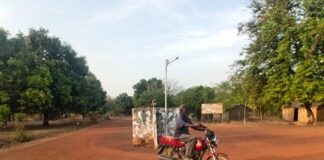 Homme conduisant une moto à la sortie de la ville de Kouki. La route est en terre à l’entrée de Kouki sur l’axe Nana-Bakassa tout ensoleillé.