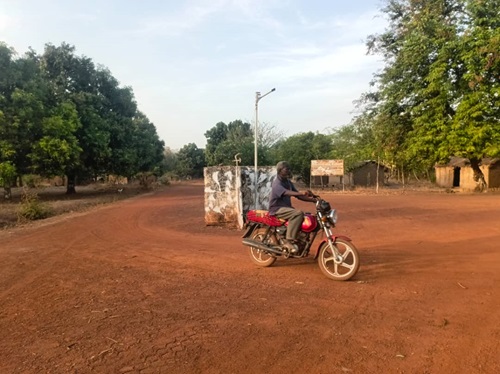 Homme conduisant une moto à la sortie de la ville de Kouki. La route est en terre à l’entrée de Kouki sur l’axe Nana-Bakassa tout ensoleillé.