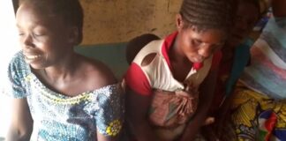 Une mère de cinq enfants souriante, vêtue d’une blouse à motifs géométriques, est assise à côté de sa sœur qui porte un enfant sur le dos avec un pagne, dans une pièce modeste.”