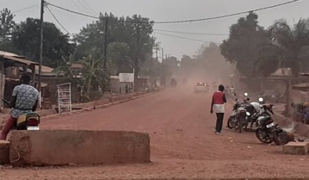 “Route poussiéreuse menant au quartier Damala avec motocyclettes et piétons sous un ciel brumeux.