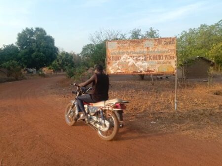 Un Homme à moto à côté d’un panneau Caritas rouillé sur une route de terre en zone rurale, affichage d’un projet communautaire à KOUKI Vita datant de 2015.”