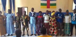 Les membres élus du bureau du comité local de gestion de plaintes de Bouar, pausant pour une photo de famille. Ils sont dans des tenues traditionnelles et décontractées posant dans une salle avec un drapeau de la République centrafricaine en arrière-plan.”
