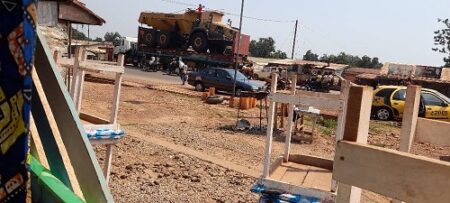Vue élargie d’un camion chargé d’un tracteur sur l’avenue de 15 mars en construction à Bangui, avec des habitants et des structures en bois.