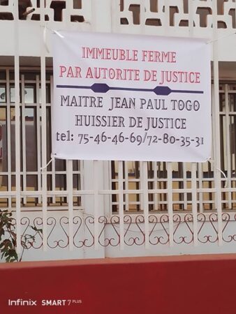 “Bannière annonçant la fermeture judiciaire d’un immeuble par Maître Jean Paul Togo, affichée sur une façade à fenêtres grillagées.” Description