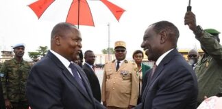 Le président du Kenya, William Ruto, serre la main du président centrafricain à Bangui, avec des dignitaires et des militaires en arrière-plan.”
