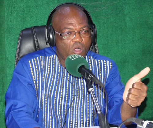 Thierry Patrick Akoloza, un ministre, est assis devant un microphone dans la Radio Ndékè - Luka, habillé en tenue traditionnelle avec des motifs dorés, faisant un geste de la main pendant qu’il parle intensément.