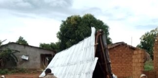 Un homme devant une maison endommagée avec un toit en tôle effondré, des résidents autour dans le village frontalier de Cantonnier. Descript