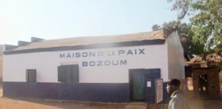 Une photo montrant l’extérieur de la “Maison de la Paix Bozoum” peinte en blanc avec une écriture noire sur le mur. Un homme marche devant le bâtiment sous un ciel clair.