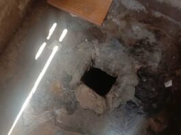 Image montrant un trou dans le sol de béton avec des débris autour, dans une pièce sombre, percé pour servir de cellule improvisée.
