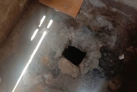 Image montrant un trou dans le sol de béton avec des débris autour, dans une pièce sombre, percé pour servir de cellule improvisée.