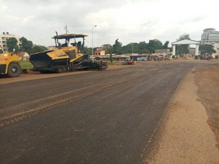 Machines de construction arrêtées sur une route partiellement goudronnée à Bangui, avec des bâtiments en arrière-plan.
