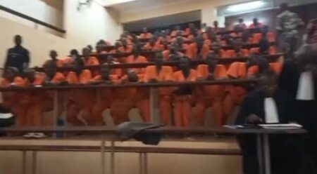 Prisonniers en tenue orange assis dans une salle d’audience lors de la première session criminelle ajournée à cause de fuites d’eau dues à la pluie, illustrant l'article sur la violence des détenus à la prison du camp de Roux