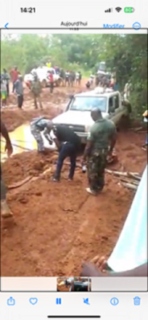 Un véhicule enlisé dans la boue sur une route dégradée en Centrafrique, entouré de commerçants et de passants tentant de dégager la route.