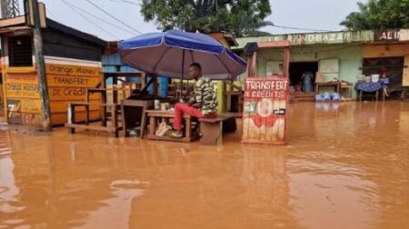 Commerçant assis sous un parasol dans une rue inondée à Bangui, devant une échoppe de transfert de crédits, après une pluie intense.