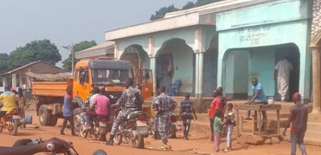 Scène de rue à Bocaranga avec un camion orange, des motos et des policiers sur des motos avec ddes piétons