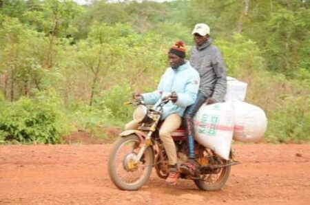 Un conducteur de moto-taxi transportant un passager et trois sacs d’arachides sur une route en terre à Bozoum. une_moto_taxi_de_bozoum