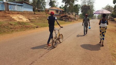 Trois personnes marchent sur une route à Mbaïki, l’une poussant un vélo, une autre tenant un sac en plastique bleu, et une femme portant un enfant avec un parapluie pour se protéger du soleil.