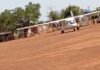 Petit avion blanc stationné sur une piste en terre rouge avec des bâtiments et des arbres en arrière-plan à l'aérodrome de BiraoPetit avion blanc des mercenaires russes stationné sur une piste en terre rouge avec des bâtiments et des arbres en arrière-plan à l'aérodrome de Birao