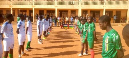 Deux équipes de football alignées sur un terrain en terre battue devant un bâtiment scolaire à Bangui"