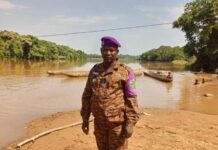 Hamidou Amine, vêtu en tenue de la douane centrafricaine, avec béret violet se tenant debout au bord de la rivière Ngamana, séparant le Cameroun avec la RCA, avec des pirogues en arrière-plan. Cet auxiliaire, recruté par le receveur des Douanes de Gamboula. CopyrightCNC