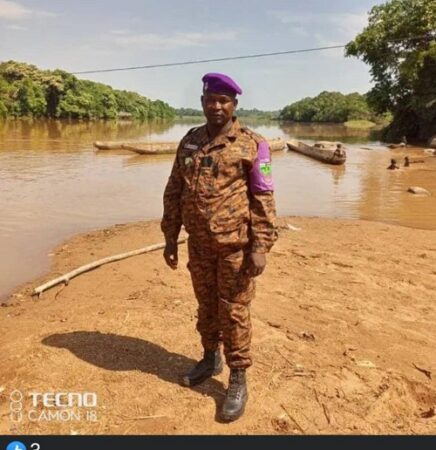 Hamidou Amine, vêtu en tenue de la douane centrafricaine, avec béret violet se tenant debout au bord de la rivière Ngamana, séparant le Cameroun avec la RCA, avec des pirogues en arrière-plan. Cet auxiliaire, recruté par le receveur des Douanes de Gamboula. CopyrightCNC