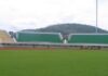 Vue des gradins et de la pelouse verte du complexe sportif Barthelemy Boganda de Bangui, avec une colline boisée en arrière-plan sous un ciel nuageux. Corbeaunews - Centrafrique