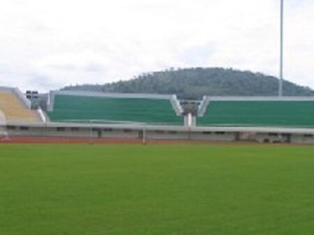 Vue des gradins et de la pelouse verte du complexe sportif Barthelemy Boganda de Bangui, avec une colline boisée en arrière-plan sous un ciel nuageux. Corbeaunews - Centrafrique