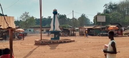 Place centrale du village Botombala, sous-préfecture de Boganda, près de la ville de Boda, dans la préfecture de la Lobaye, avec un monument, des boutiques, et habitants sous un ciel clair, après des attaques armées.