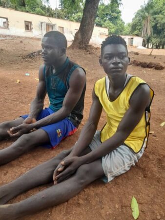 Deux jeunes miliciens Azandés assis par terre, interpellés par des mercenaires russes à Zemio après avoir tué trois éleveurs Peuls à 8 kilomètres de la ville dans la brousse."