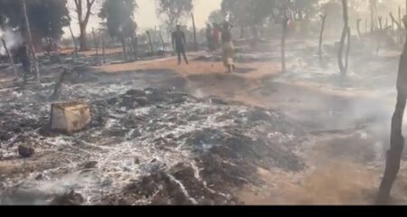 Vue panoramique des ruines du village de Wadjia après un incendie. Le sol est couvert de cendres, des arbres brûlés et des structures en bois détruites sont visibles, tandis que des habitants errent parmi les décombres.