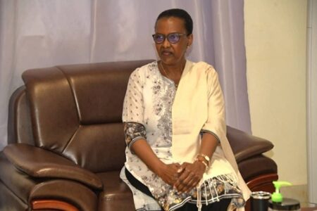 Madame Valentine Rugwabiza, représentante du Secrétaire général des nations-unies en Centrafrique, assise sur un canapé en cuir marron au palais de la renaissance en face du Président Touadera, avec un châle clair sur l’épaule. Elle porte des lunettes et une tunique blanche ornée de motifs floraux gris et noirs. Elle a les mains croisées sur les genoux et regarde légèrement sur le côté. À droite, on peut regarder aisément une bouteille de gel désinfectant vert sur une table.